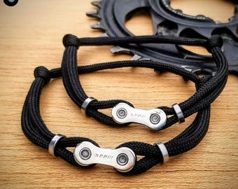 Puede incluir: Dos pulseras de paracord negro con eslabones de cadena de bicicleta plateados. Los eslabones de la cadena están grabados con la palabra "SRAM".