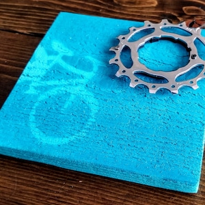 Puede incluir: Un posavasos cuadrado de madera azul turquesa con un piñón de bicicleta plateado en la parte superior.