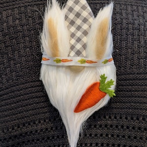 Puede incluir: Un gnomo blanco con una larga barba blanca y un gorro a cuadros gris y blanco. El gnomo sostiene una zanahoria de fieltro con hojas verdes.