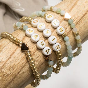 Könnte beinhalten: Vier Armbänder mit Perlen mit goldenen und weißen Akzenten. Eines buchstabiert "MAMA" mit goldenen Buchstaben auf weißen Perlen. Ein anderes buchstabiert "DEVIN". Jedes Armband hat einen Herzanhänger. Die Armbänder sind auf einem Holzstab ausgestellt.