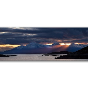 Puede incluir: Una vista panorámica de una cordillera con picos cubiertos de nieve, iluminada por una espectacular puesta de sol. Los rayos del sol atraviesan nubes oscuras, proyectando un brillo dorado sobre el paisaje. Las montañas se reflejan en las aguas tranquilas de un lago o una bahía.
