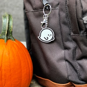 May include: A white ghost keyring with a black outline and a small, friendly face. The keyring is attached to a silver chain and is hanging from a dark grey rucksack.