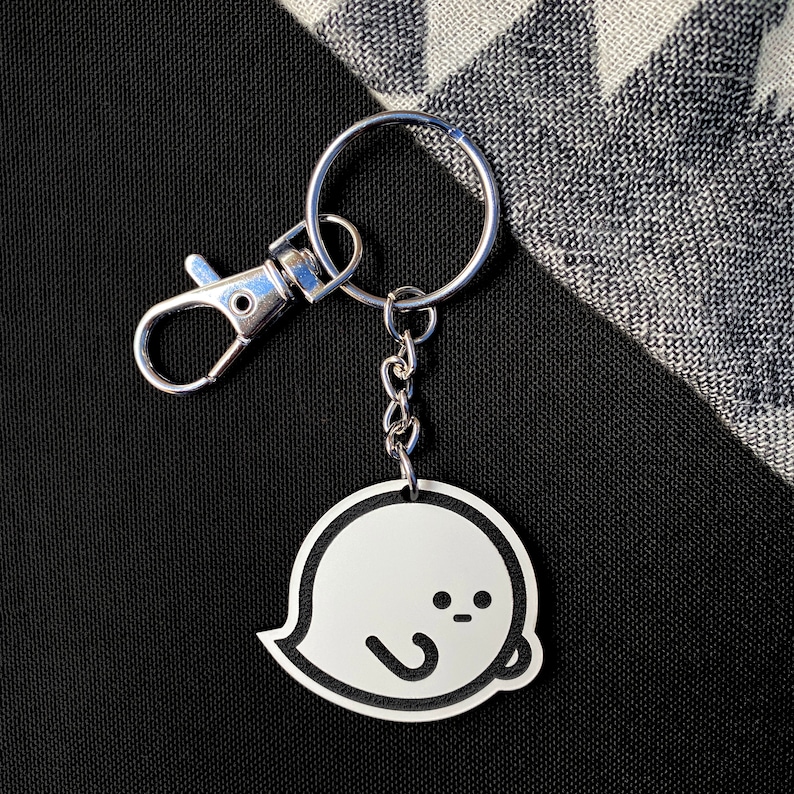 May include: A silver keyring with a white ghost charm. The ghost has a simple, cartoon-like face with two black eyes and a small mouth.
