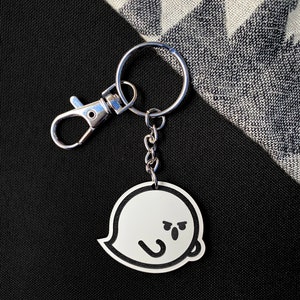 May include: A silver keyring with a black and white ghost charm. The ghost has a frowning face with two small eyes and a mouth.