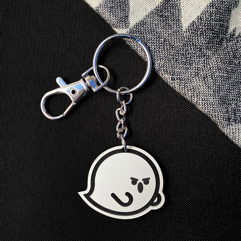 May include: A silver keyring with a black and white ghost charm. The ghost has a frowning face with two small eyes and a mouth.