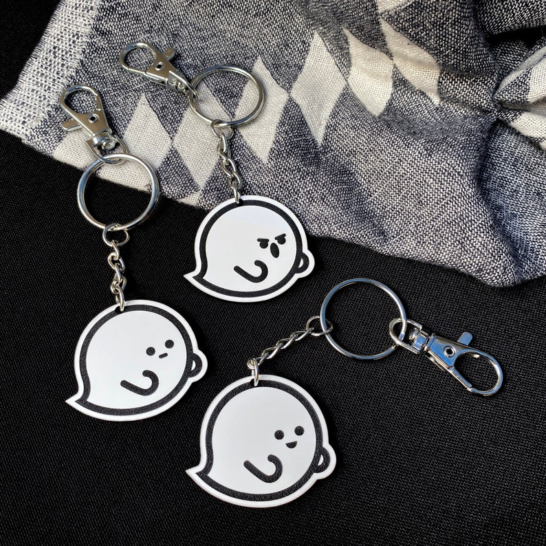 May include: Three white ghost keychains with black outlines and different facial expressions. The keychains are attached to silver key rings.