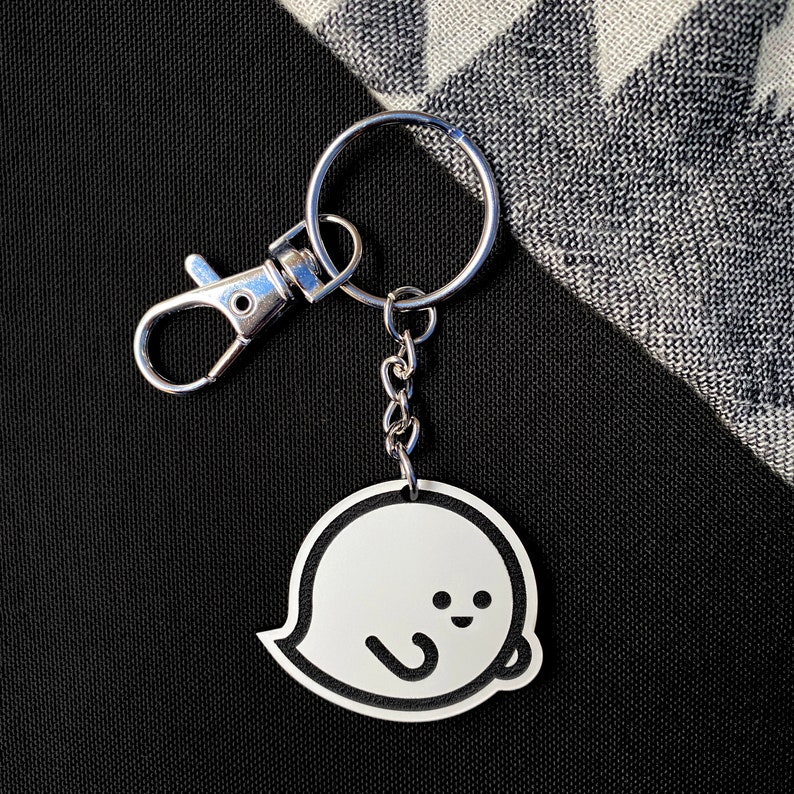 May include: A silver keyring with a white ghost charm. The ghost has a simple, smiling face and a small, curved arm.