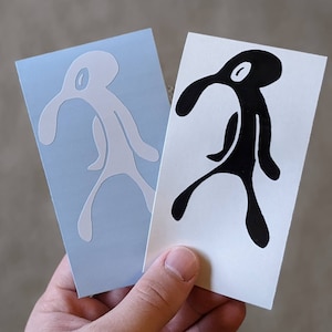 May include: Two stickers featuring a cartoon character with a long nose and a single eye. One sticker is white on a blue background, and the other is black on a white background.
