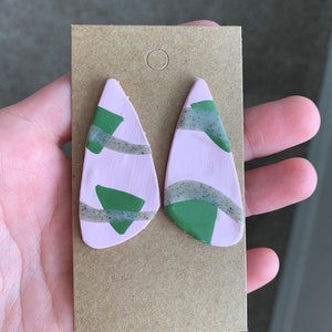 May include: A pair of pink and green geometric dangle earrings. The earrings are made of polymer clay and have a unique abstract design.