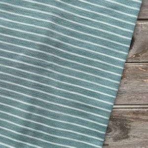 May include: Close-up of a fabric with a teal and white striped pattern. The fabric is angled, with the stripes running diagonally across the frame. The fabric is on a wooden surface.