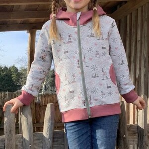 Könnte beinhalten: Ein hellgrauer Kapuzenpullover mit Reißverschluss und einem wiederkehrenden Muster aus kleinen Hasen, Schlittschuhen und Schneeflocken in Rosa und Grau. Das Kleidungsstück hat eine einfarbige rosa Kapuze, rosa Ärmelbündchen und zwei Taschen.