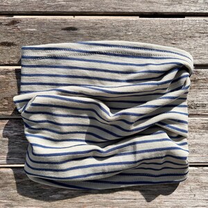 May include: A folded, tubular neck gaiter made of a soft, light-colored fabric with horizontal navy blue stripes. The fabric is gathered and twisted, showing the interior and exterior of the tube. The item is laying on a wooden surface.