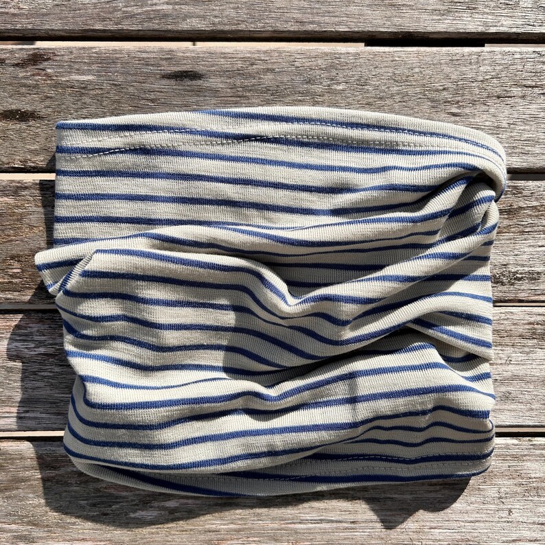 May include: A folded, tubular neck gaiter made of a soft, light-colored fabric with horizontal navy blue stripes. The fabric is gathered and twisted, showing the interior and exterior of the tube. The item is laying on a wooden surface.