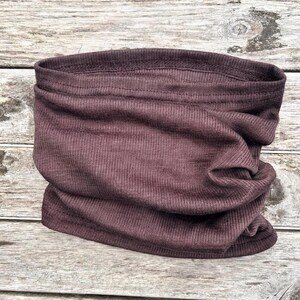 May include: A dark mauve-colored neck gaiter made of ribbed knit fabric. The tube-shaped accessory is designed to be worn around the neck and can be pulled up over the mouth and nose. The fabric is soft and appears to be stretchy.