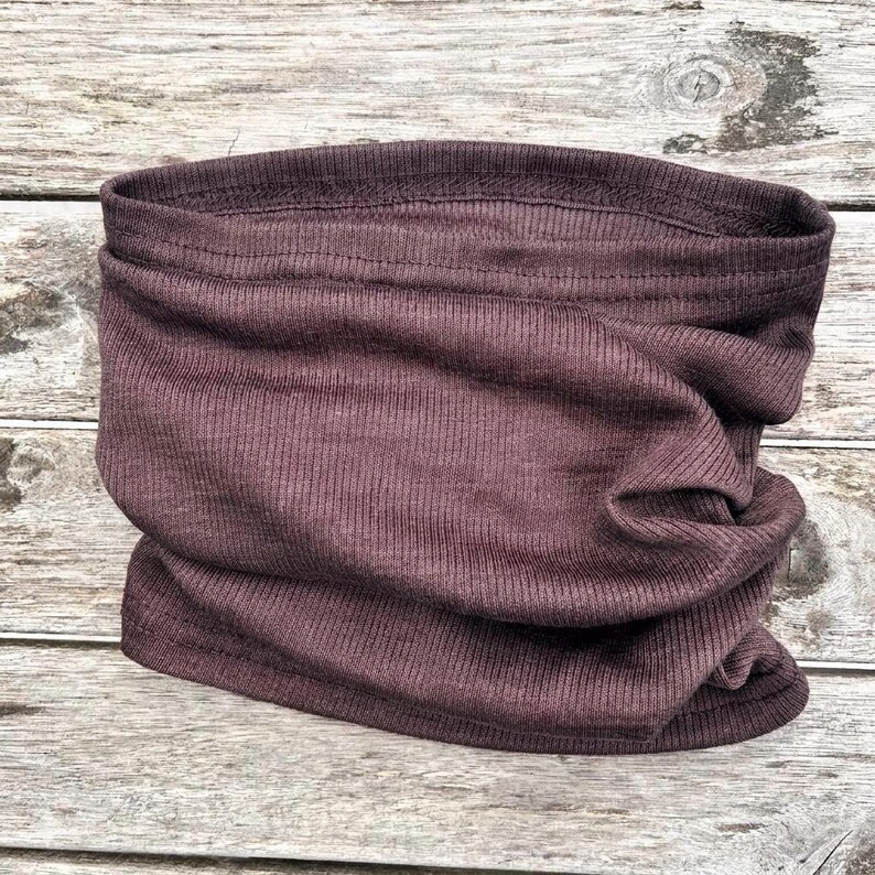 May include: A dark mauve-colored neck gaiter made of ribbed knit fabric. The tube-shaped accessory is designed to be worn around the neck and can be pulled up over the mouth and nose. The fabric is soft and appears to be stretchy.