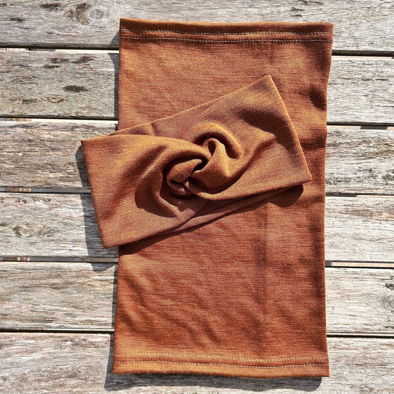 May include: A rust-colored neck gaiter and matching twisted headband are displayed on a wooden surface. The fabric has a subtle texture, and the headband is knotted at the top. The items appear to be made of a soft, comfortable material.