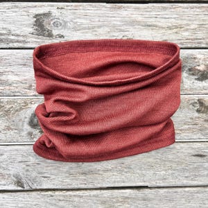 May include: A rust-colored neck gaiter made of a ribbed knit fabric. The tube-shaped scarf is folded and draped, showcasing its texture and form. The fabric appears soft and flexible, suitable for various weather conditions.
