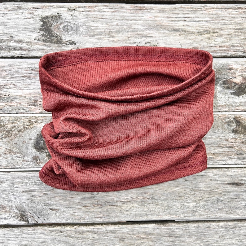May include: A rust-colored neck gaiter made of a ribbed knit fabric. The tube-shaped scarf is folded and draped, showcasing its texture and form. The fabric appears soft and flexible, suitable for various weather conditions.