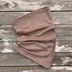 May include: A folded, taupe-colored neck gaiter made of a ribbed knit fabric. The fabric has a subtle striped pattern and is sewn with a matching thread. The neck warmer is displayed on a weathered, gray wooden surface.