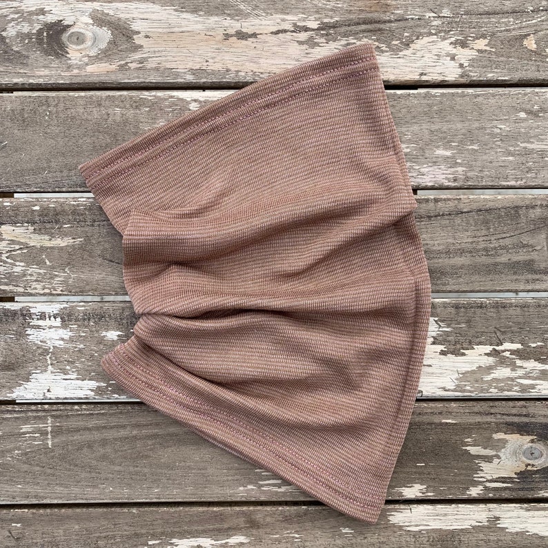 May include: A folded, taupe-colored neck gaiter made of a ribbed knit fabric. The fabric has a subtle striped pattern and is sewn with a matching thread. The neck warmer is displayed on a weathered, gray wooden surface.