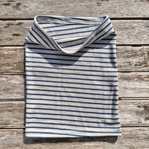 May include: A folded, tubular neck gaiter with horizontal stripes in navy blue and off-white. The fabric is soft and appears to be a knit material. The neck warmer is laying on a wooden surface.