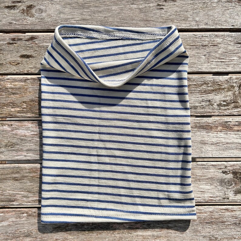 May include: A folded, tubular neck gaiter with horizontal stripes in navy blue and off-white. The fabric is soft and appears to be a knit material. The neck warmer is laying on a wooden surface.