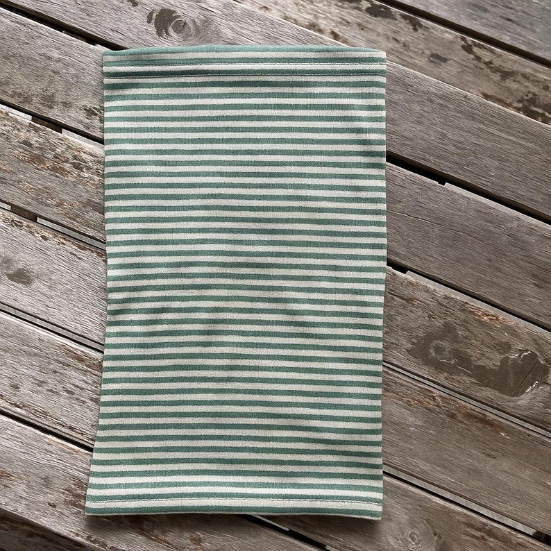 May include: A green and white striped neck gaiter, a tube-shaped accessory. The fabric has horizontal stripes of light green and white. The gaiter is laying on a weathered wooden surface.