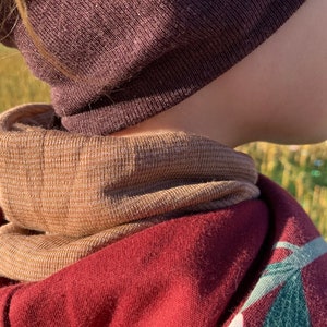May include: A close-up of a person wearing a brown beanie and a tan and brown neck warmer. The neck warmer is layered over a maroon hooded sweatshirt with a green and white design. The background is blurred.