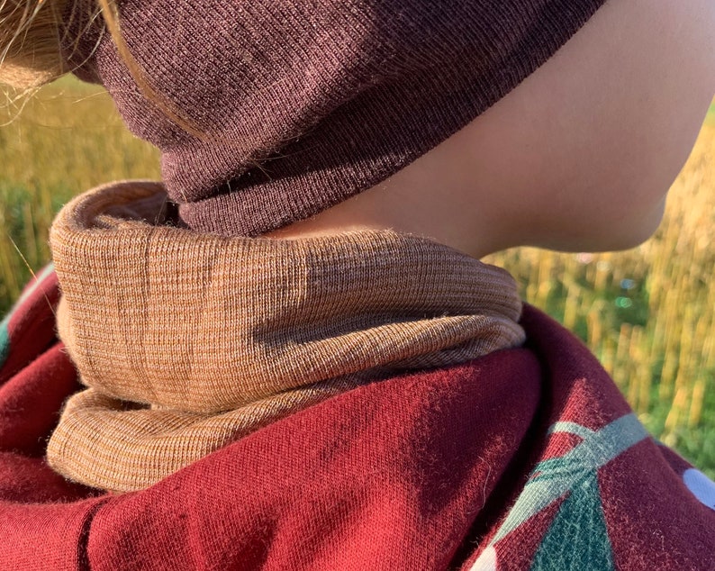 May include: A close-up of a person wearing a brown beanie and a tan and brown neck warmer. The neck warmer is layered over a maroon hooded sweatshirt with a green and white design. The background is blurred.