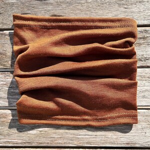 May include: A folded, rust-colored neck gaiter made of soft fabric. The tube scarf is designed to be worn around the neck and face for warmth and protection. The fabric has a ribbed texture and is stitched at the top and bottom.