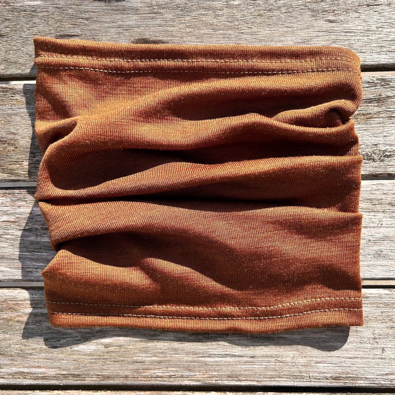May include: A folded, rust-colored neck gaiter made of soft fabric. The tube scarf is designed to be worn around the neck and face for warmth and protection. The fabric has a ribbed texture and is stitched at the top and bottom.