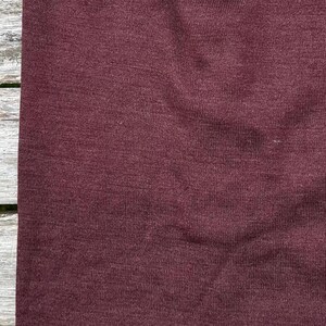 May include: Close-up of a burgundy fabric with a textured surface. The fabric appears to be a knit or woven material, with subtle variations in color and texture. The color is a deep, rich shade of red-violet.