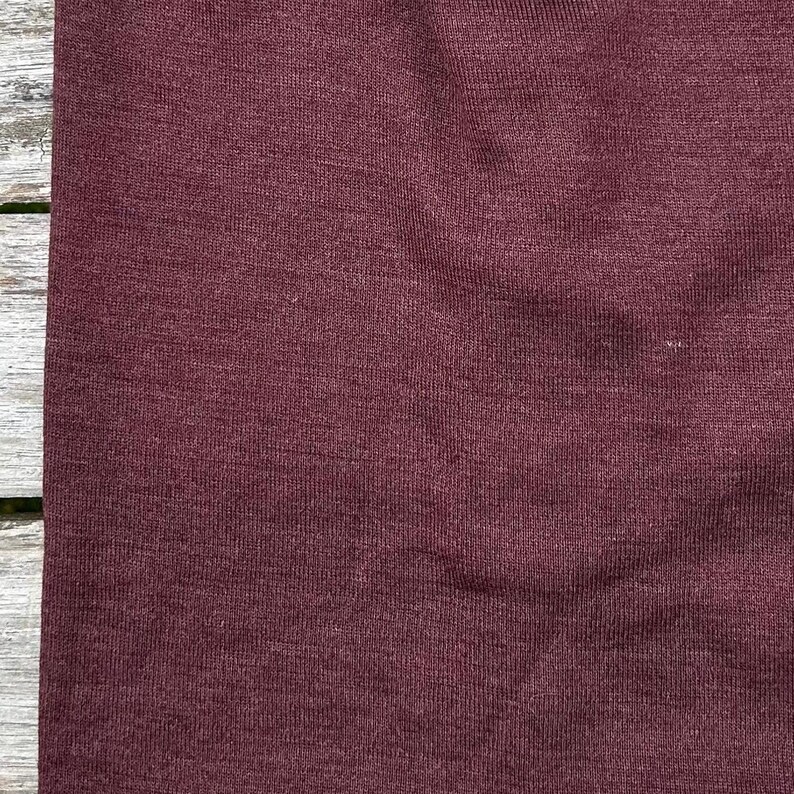 May include: Close-up of a burgundy fabric with a textured surface. The fabric appears to be a knit or woven material, with subtle variations in color and texture. The color is a deep, rich shade of red-violet.