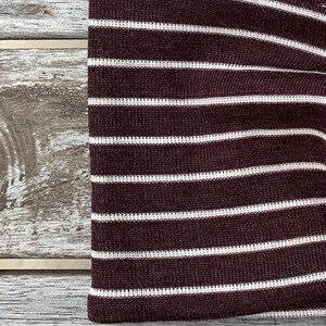 May include: Close-up of a maroon and white striped fabric. The fabric has horizontal white stripes against a maroon background. The fabric is folded over, showing the texture and pattern of the material. The fabric is likely part of a garment.