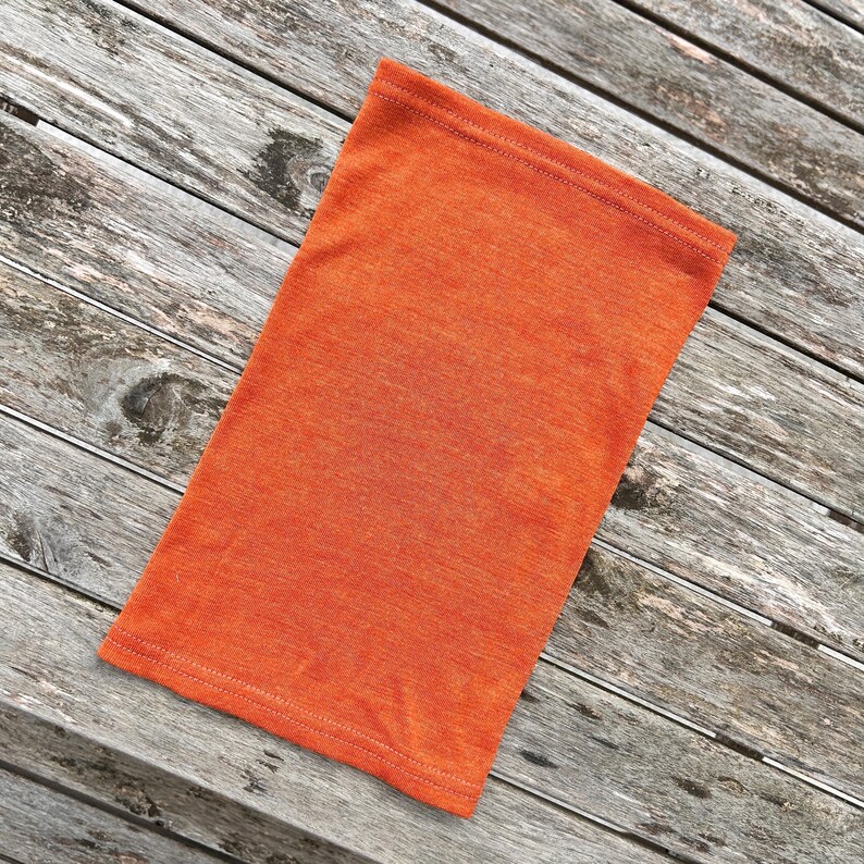 May include: An orange neck gaiter made of a soft, textured fabric. The rectangular tube is folded in half and lies on a weathered wooden surface. The seams are visible along the top and bottom edges.