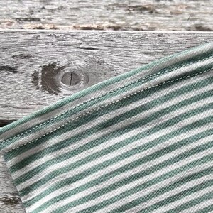 May include: Close-up of a section of a striped fabric. The fabric features alternating stripes of white and a muted green color. The edges are finished with a double-stitched seam in a matching green thread. The background is a weathered wooden surface.