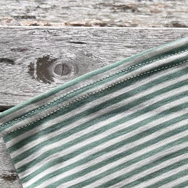 May include: Close-up of a section of a striped fabric. The fabric features alternating stripes of white and a muted green color. The edges are finished with a double-stitched seam in a matching green thread. The background is a weathered wooden surface.