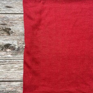 May include: A close-up of a solid red fabric, likely a piece of clothing, against a backdrop of weathered, gray wooden planks. The fabric's texture is visible, and a small dark spot is present.