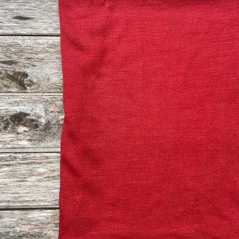 May include: A close-up of a solid red fabric, likely a piece of clothing, against a backdrop of weathered, gray wooden planks. The fabric's texture is visible, and a small dark spot is present.