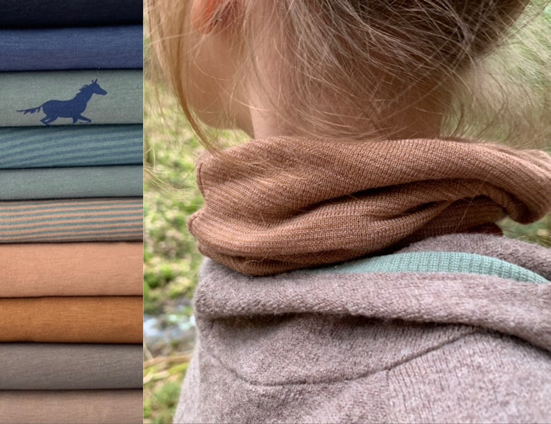 May include: A stack of folded fabric in various shades of blue, green, and tan. One shirt has a blue horse graphic. A person wears a brown ribbed neck warmer and a gray hooded sweatshirt.