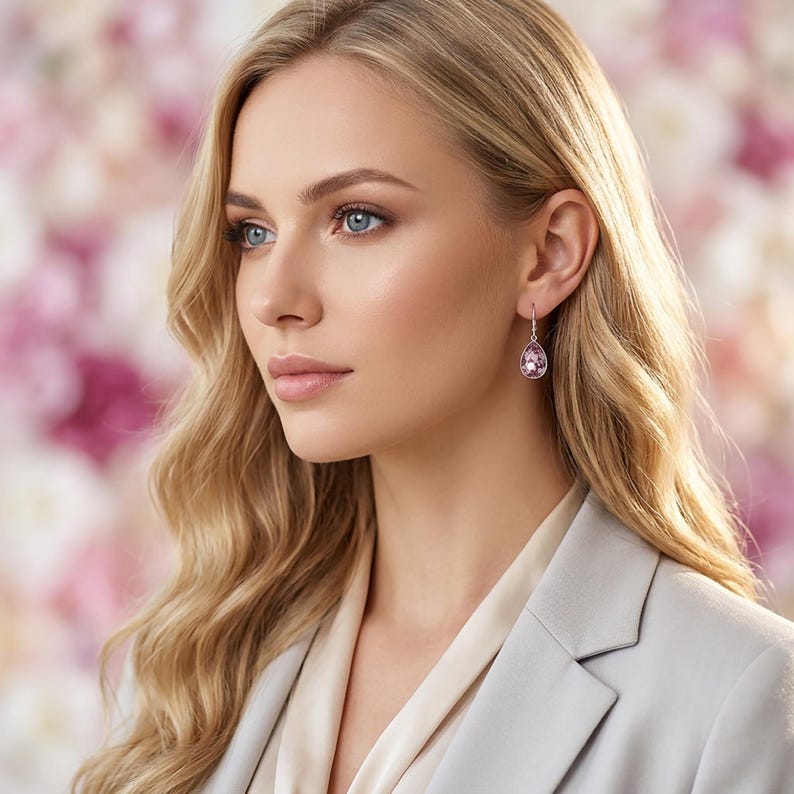 May include: A woman wearing a pair of teardrop-shaped earrings with a pink gemstone. The earrings are set in a silver-colored metal and hang from a small hook. The woman is wearing a light gray blazer and a cream-colored blouse.