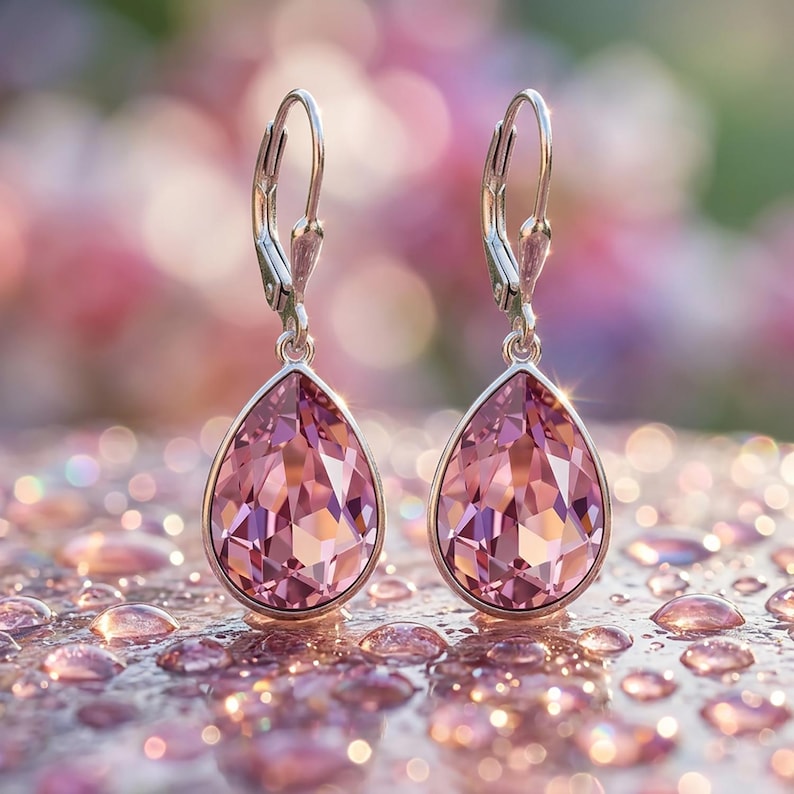 May include: A pair of elegant drop earrings with a pear-shaped, faceted pink crystal. The crystals are set in a silver-colored metal and feature a lever-back closure. The earrings are displayed on a reflective surface.