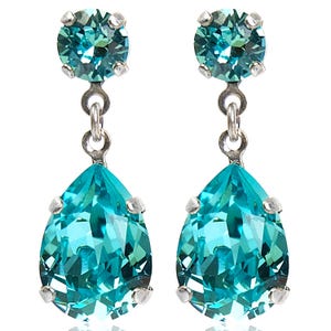 Könnte beinhalten: Ein Paar elegante Ohrhänger mit tropfenförmigen, facettierten Kristallen in Türkis. Jeder Ohrring hat einen kleineren, runden Kristall oben, verbunden durch eine silberfarbene Metallkette. Die Kristalle sind in silberfarbenem Metall gefasst.
