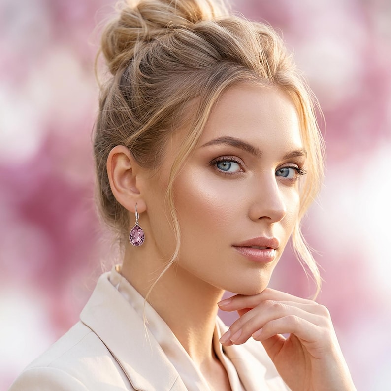 May include: A woman wearing a light pink blazer and a pair of drop earrings. The earrings feature a pear-shaped, light purple gemstone. The background is a soft pink and white.