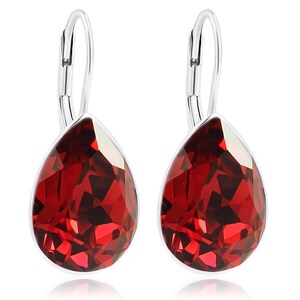 Könnte beinhalten: Ein Paar silberfarbene Ohrhänger mit großen, facettierten, tropfenförmigen roten Kristallen. Die Kristalle sind in eine silberne Lünette gefasst und haben eine reflektierende, facettenreiche Oberfläche. Die Ohrringe haben einen Bügelverschluss.
