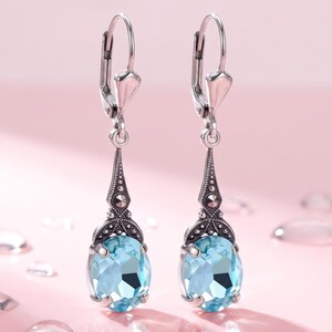 Könnte beinhalten: Ein Paar silberne Ohrhänger mit hellblauen, oval geschliffenen Edelsteinen. Jeder Ohrring verfügt über eine dekorative Silberfassung mit filigranen Metallarbeiten und einem Klappbügelverschluss. Die Steine sind facettiert und klar.