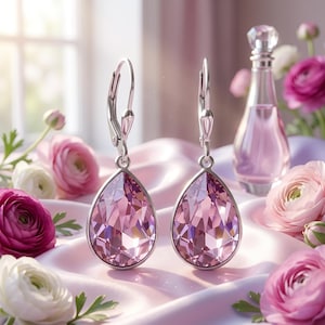 May include: A pair of silver drop earrings with large, faceted, teardrop-shaped pink crystals. The earrings are displayed on a pink satin surface, surrounded by pink and white flowers and a perfume bottle.