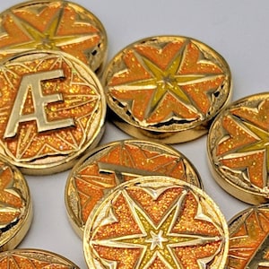 Könnte beinhalten: Nahaufnahme von goldfarbenen Spielsteinen mit einem Sternmuster und dem Buchstaben "A" auf einigen. Die Spielsteine haben eine orangefarbene Glitzer-Oberfläche.