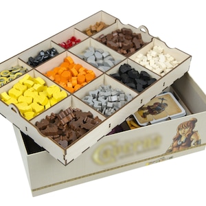 May include: A wooden organizer with compartments for storing game pieces. The organizer is filled with various colored wooden pieces, including red, orange, yellow, brown, gray, black, and white. The organizer is sitting on top of a game box with a woman's face on the front.