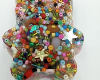Op de afbeelding: Een kleurrijk teddybeerbeeldje van hars, gevuld met een assortiment kralen in verschillende kleuren, waaronder rood, geel, blauw en groen. De beer heeft een gouden ster en een zilveren ster ingebed in de hars.